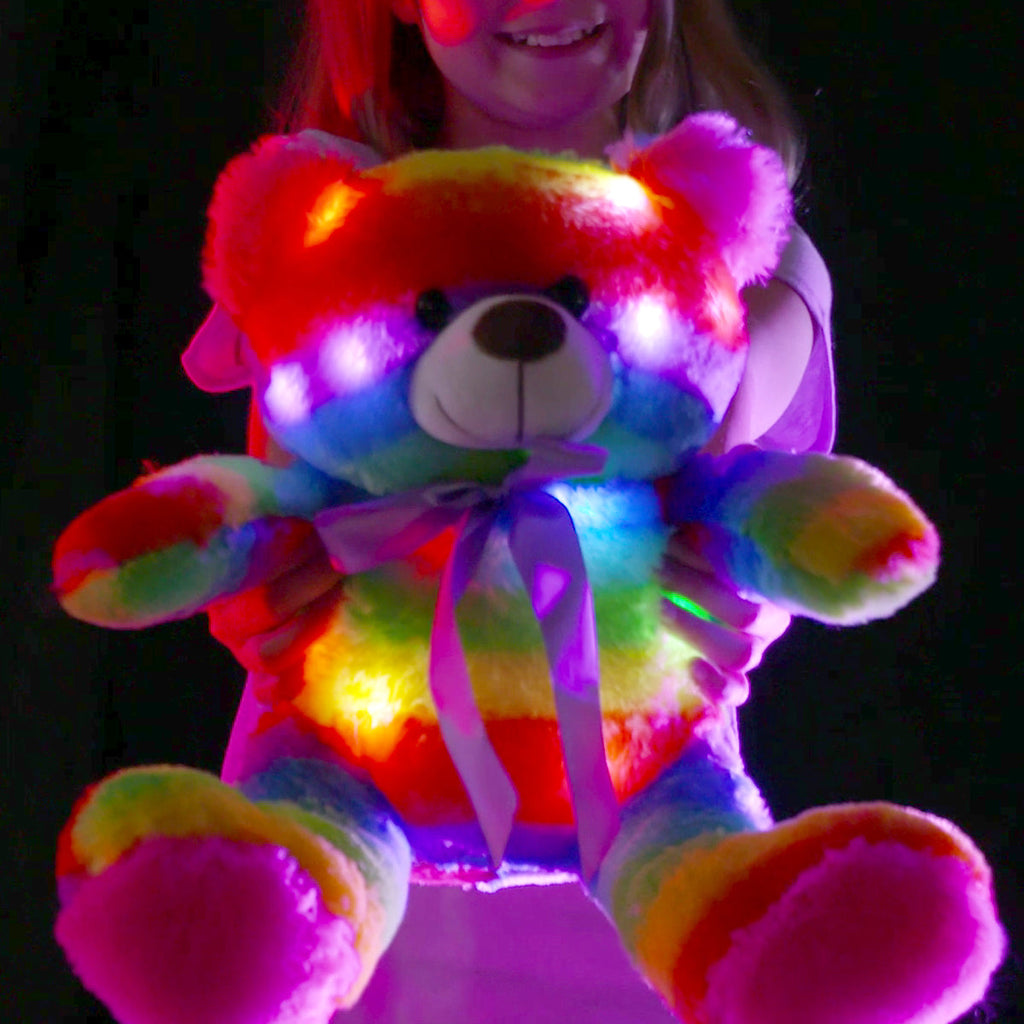 Rainbow teddy bear glowing in the dark held by a child with a joyful expression.