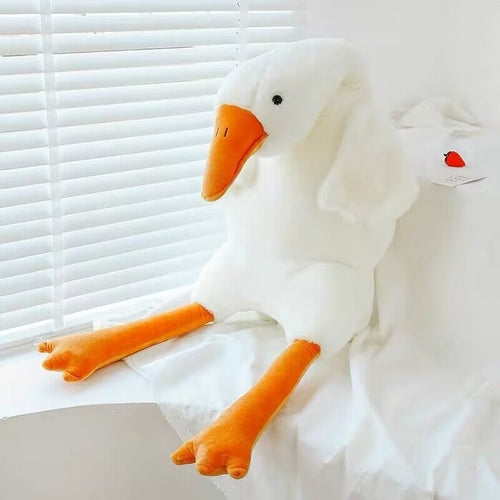 White goose plush seated beside a window with orange feet extended